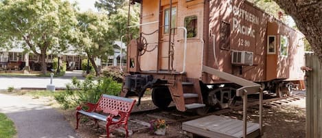 Un vagón de tren antiguo con un banco y una pequeña mesa frente a él, en un área arbolada con un sendero pavimentado.