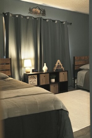 A bedroom with two beds, a nightstand with a lamp, a clock, and a decorative sign. - Charming 2-bedroom house in Wasilla perfect for your getaway (Wasilla)