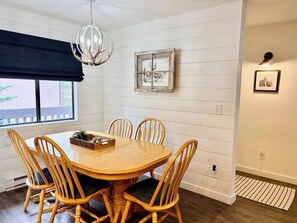 A dining area with a round wooden table and six chairs, a chandelier, a window with a dark valance, and a framed picture on the wall.