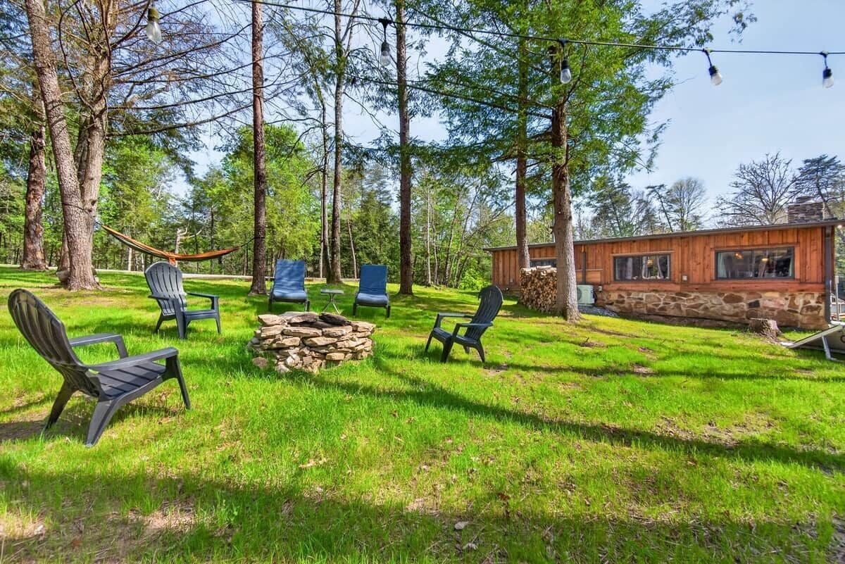 A wooden cabin with a stone exterior, surrounded by a grassy yard with several Adirondack chairs and a hammock.