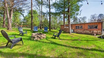 A wooden cabin with a stone exterior, surrounded by a grassy yard with several Adirondack chairs and a hammock.