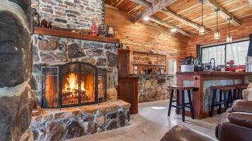 A stone fireplace with a fire, wooden walls, a bar area with stools, and a large window.