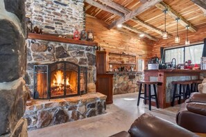 A stone fireplace with a fire, wooden walls, a bar area with stools, and a large window. - Stagecoach Saloon Riverside cabin with stone pizza oven (Cumberland County)