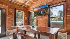 Un interior de madera con una mesa grande y bancos, ventanas amplias y un cuadro en la pared.