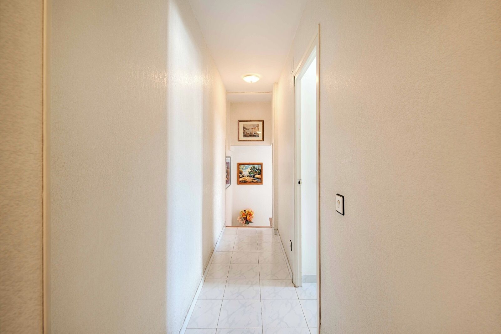 A hallway with white walls and a tiled floor, leading to a room with a door and a framed picture on the wall.