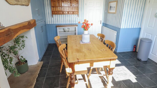 Un comedor con una mesa de madera, sillas, un florero con flores, una zona de cocina con paredes a rayas azules y una puerta blanca.