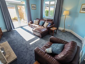 A living room with a brown leather sofa, a wooden coffee table, and a lamp. There is a window with curtains and a view of the outside. - Sorrento (Mablethorpe)
