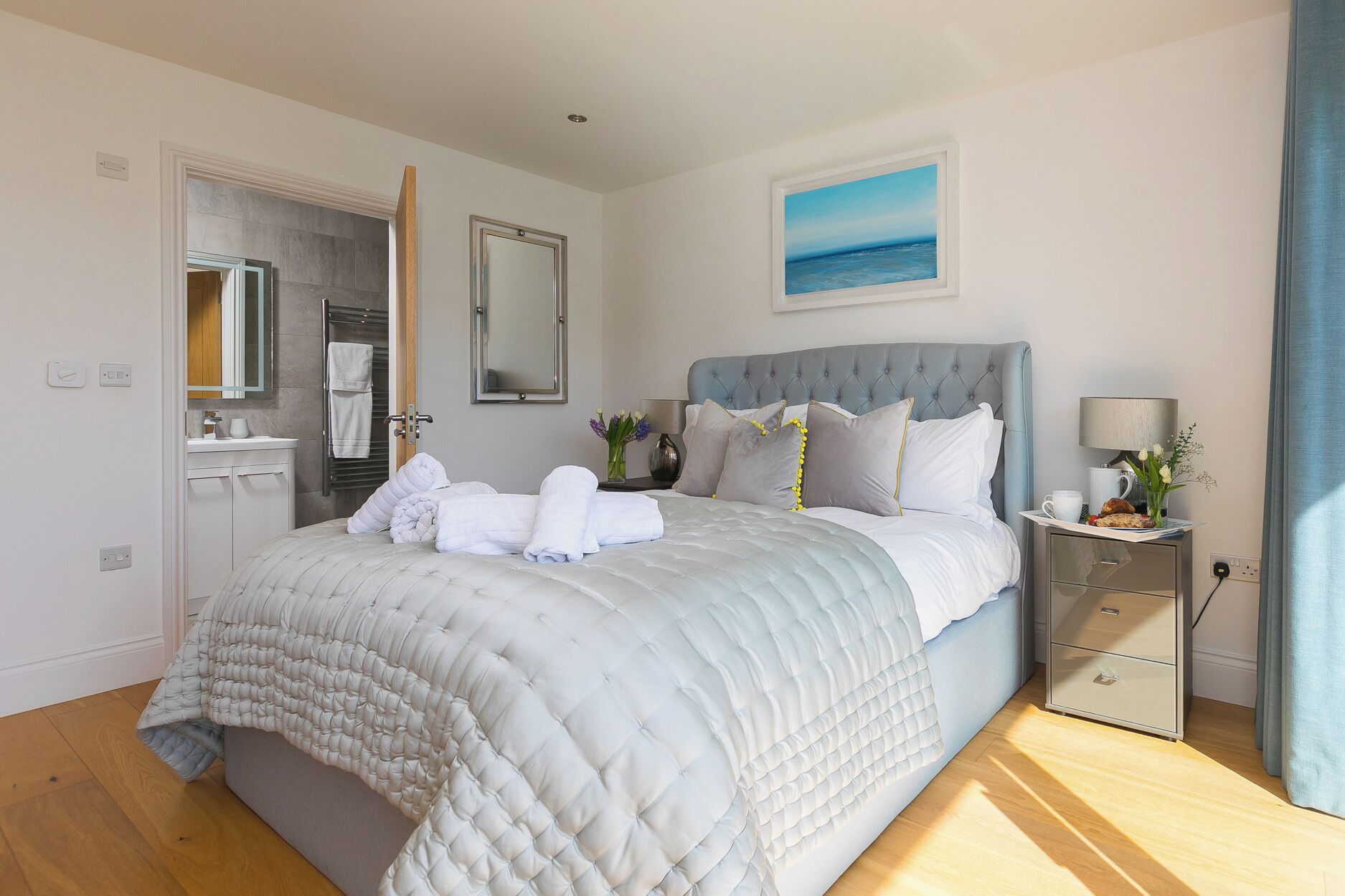 Une chambre à coucher avec un lit, une table de chevet, un miroir, un tableau, et une salle de bain visible à travers une porte ouverte.