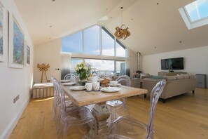 A dining area with a wooden table and clear chairs, a living room with a sofa and TV, and large windows offering a view. - 2 Remera (St Ives)