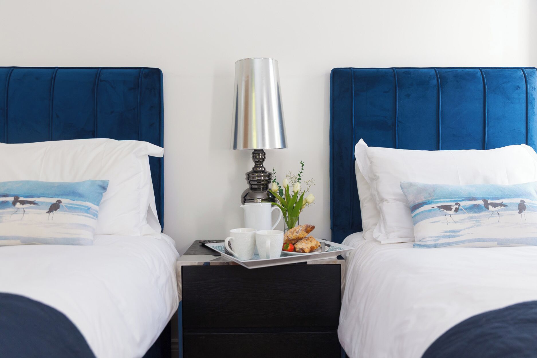 A hotel room with two beds, a nightstand with a lamp, a tray with breakfast items, and a vase with flowers.