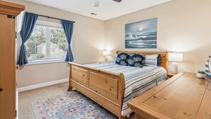 A bedroom with a wooden bed, a ceiling fan, a window with curtains, a painting on the wall, and a small table with a plant. - Pecan Pointe Cottage - Pet Friendly (Brunswick)