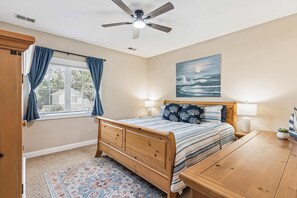 A bedroom with a wooden bed, a ceiling fan, a window with curtains, a painting on the wall, and a small table with a plant. - Pecan Pointe Cottage - Pet Friendly (Brunswick)