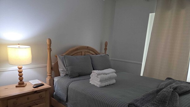 Una cama bien hecha con colcha gris y almohadas, una mesita de noche con lámpara y una mesita auxiliar de madera con control remoto.
