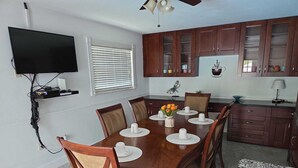 Un comedor con una mesa de madera preparada para cuatro, un televisor montado en la pared y una cocina con armarios de madera.