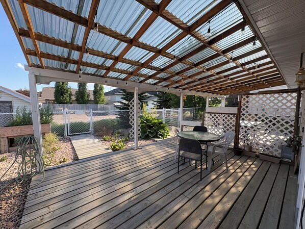 A covered patio with a wooden deck, a round table with chairs, and a metal fence in the background. - Mama’s House of Relaxation (Sundre)