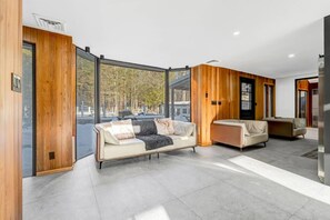 A modern interior with a sofa and armchairs, wooden paneling, and a large window offering a view of trees.