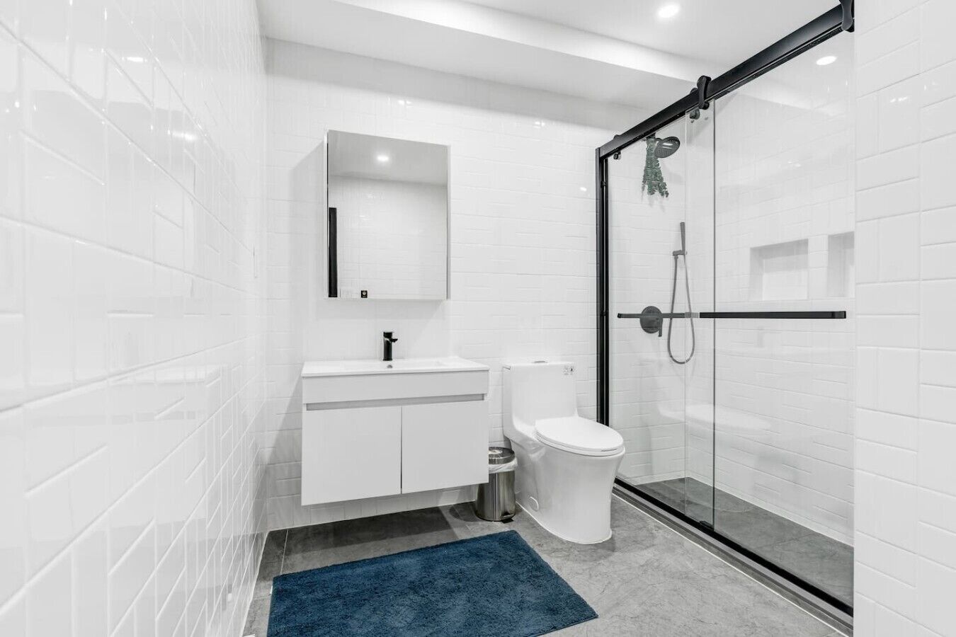 A modern bathroom with white tiles, a glass shower enclosure, a white toilet, and a blue rug.