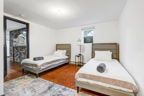 A room with two single beds, a small table, a lamp, and a view of a brick building through a glass door.