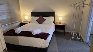 Un lit bien fait, recouvert d’un plaid marron, avec deux lampes de chevet, une table de chevet en bois et un porte-manteau en métal.