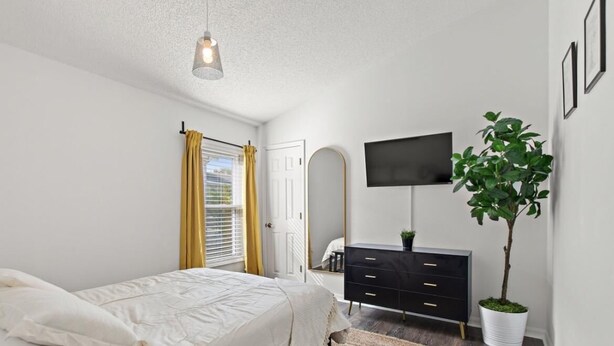 Ein Schlafzimmer mit einem Bett, einem Schminktisch, einem Fernseher, einer Pflanze und einem Fenster mit Vorhängen.