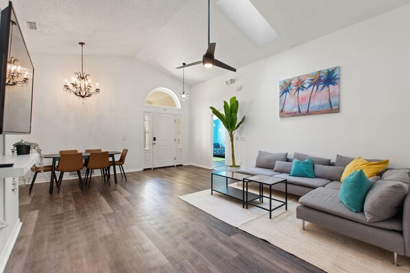 A modern living room with a grey sofa, a dining table with chairs, a chandelier, a ceiling fan, and a painting of palm trees. - Amazing 3/2 vacation home with private pool!! (Davenport)