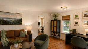 A living room with a brown sofa, a green armchair, a black coffee table, and a dining area in the background. - Couples King Bed Cottage with Fire Pit & Swing, Near Asheville & Biltmore in WNC (Leicester)