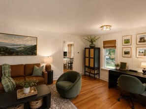 Ein Wohnzimmer mit braunem Sofa, einem grünen Sessel, einem schwarzen Couchtisch und einem Essbereich im Hintergrund.
