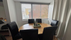 Een ronde tafel met zes stoelen, een raam met zonwering en een gordijn.