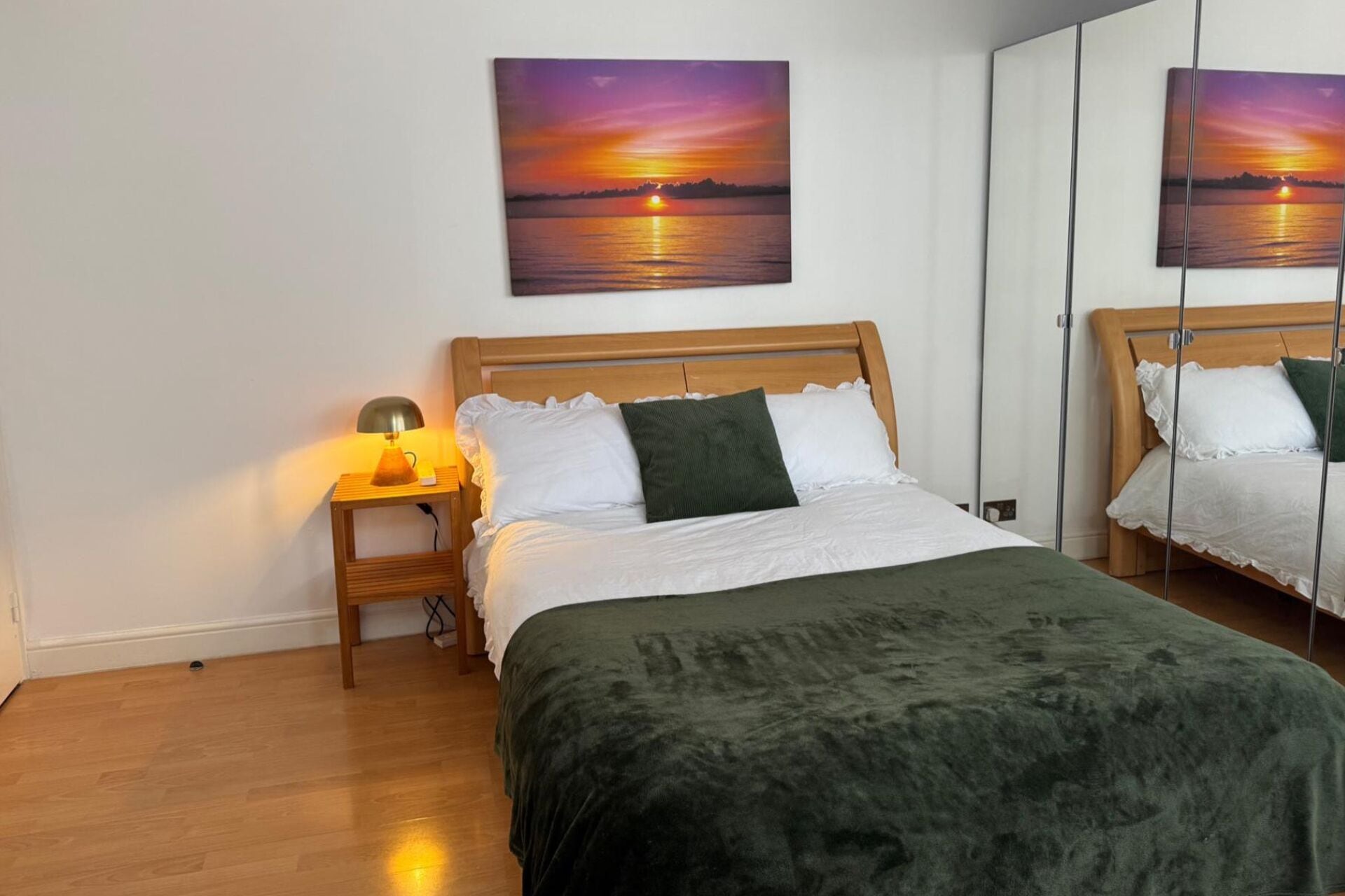 Een slaapkamer met een houten bed, wit beddengoed, een groene plaid, een bedlampje en een schilderij van een zonsondergang aan de muur.