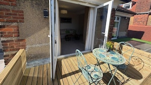 Une terrasse en bois avec une table et des chaises, une porte vitrée donnant sur une pièce avec un canapé et une table à manger, et un mur en briques.