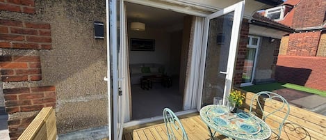 En træterrasse med et bord og stole, en glasdør ind til et rum med en sofa og et spisebord, samt en murstensvæg.