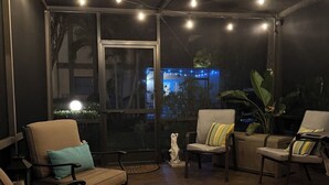 Een overdekt terras met stoelen en een standbeeld, grote ramen en lichtslingers.