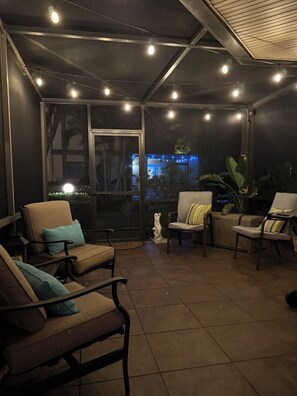 A covered outdoor seating area with chairs and a statue, large windows, and string lights. - Coastal Escape Near Sanibel, Pool, Lanai & Beach Adventures - Tropical Tides Townhome - Roelens (Fort Myers)