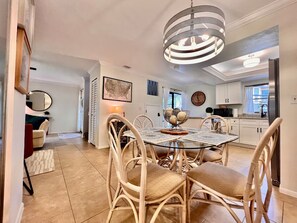 Un coin repas avec une table en verre, des chaises en osier, un luminaire suspendu et une cuisine en arrière-plan.