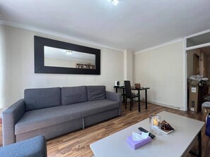 Ein Wohnzimmer mit grauem Sofa, einem Esstisch mit Stühlen, einem kleinen Tisch mit Taschentuchbox und Fernbedienung sowie einem Spiegel an der Wand.