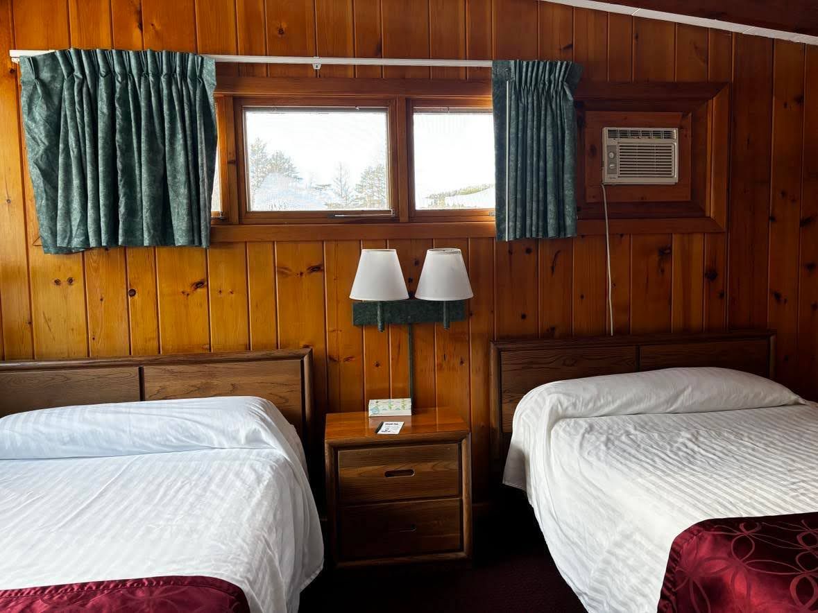 Ein Zimmer mit Holzwänden, zwei Betten, einem Nachttisch mit Lampe und einem mit Vorhängen versehenen Fenster.