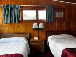 Ein Zimmer mit Holzwänden, zwei Betten, einem Nachttisch mit Lampe und einem mit Vorhängen versehenen Fenster.