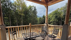 A covered outdoor deck with wooden railings, a table, and chairs. - Luxury Stowe Home w/ Sauna & Hot Tub (Stowe)