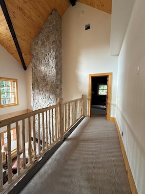 Un pasillo amplio con pared de piedra, barandilla de madera y suelo alfombrado que conduce a una habitación.