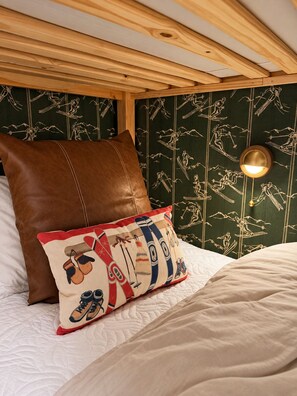 A bed with a quilted white cover, a brown leather pillow, and a colorful pillow with ski and snowboard designs.