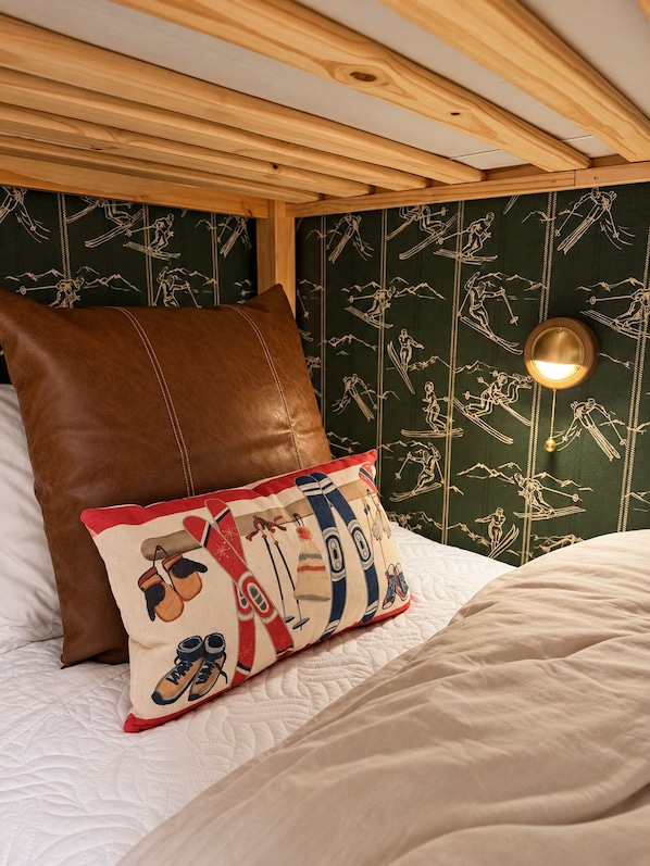 Una cama con cubrecolcha blanco acolchado, almohada de cuero marrón y almohada colorida con diseños de esquí y snowboard.
