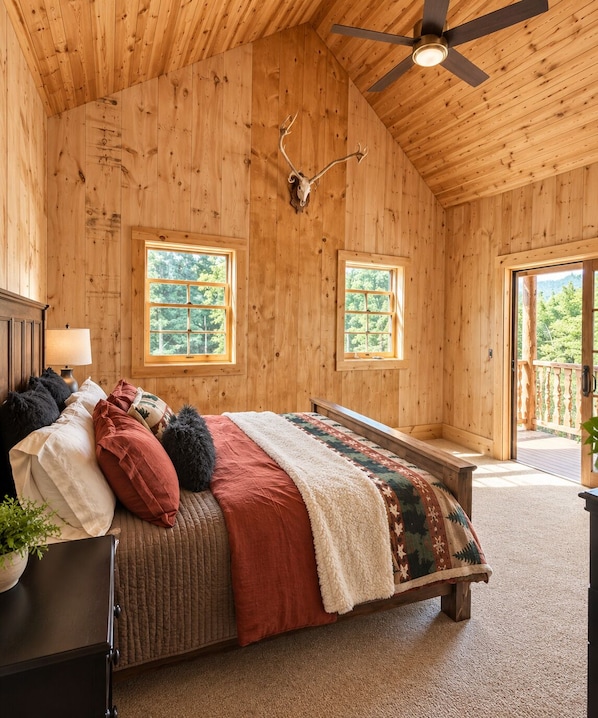 Un dormitorio de cabaña de madera con una cama, dos ventanas, un ventilador de techo y una decoración de cráneo de ciervo en la pared.