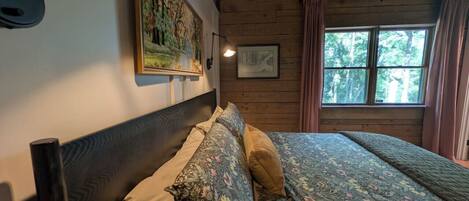 Une pièce avec un plafond en bois, un lit avec une literie à motifs floraux, une tête de lit et une fenêtre avec des rideaux.