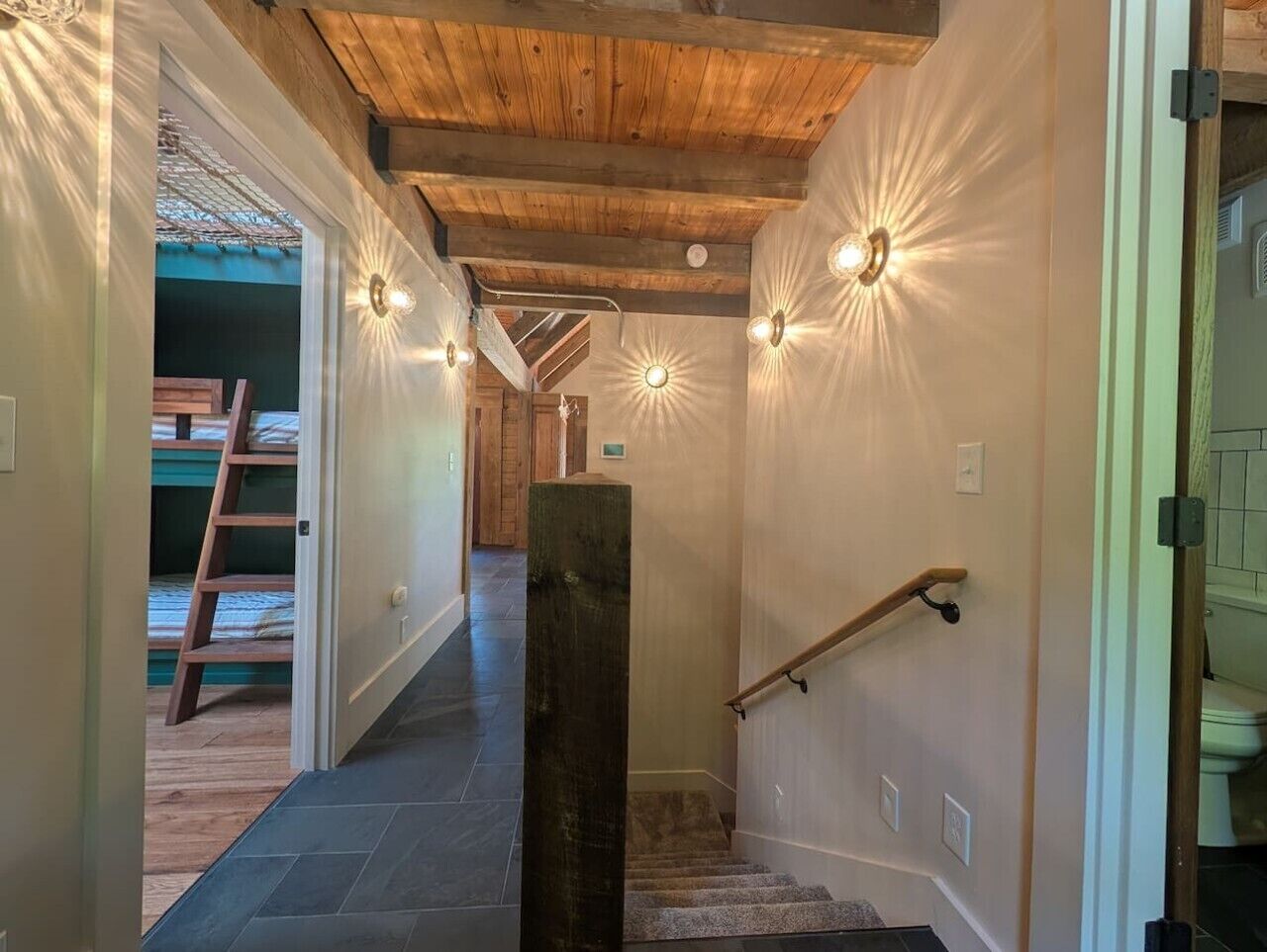 A staircase with wooden railings and wall-mounted lights, leading to a room with a visible bed and a ladder.