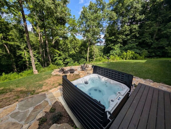 A hot tub in a backyard with a stone patio, surrounded by trees and a wooden deck. - Heated Indoor Pool | Net Bed | Theater | 100 acres (Stanton)