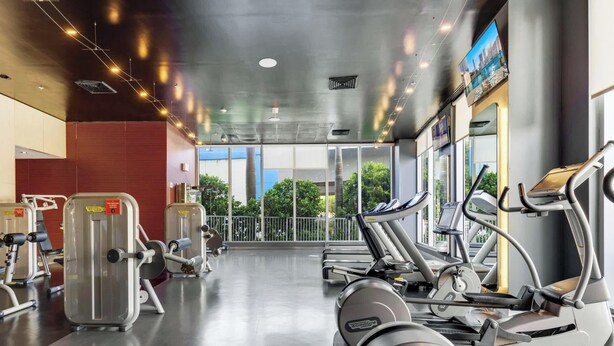 A gym with cardio machines, weight machines, and a large window with a view of greenery. - High-Floor Studio with Intracoastal Views (Hallandale Beach)