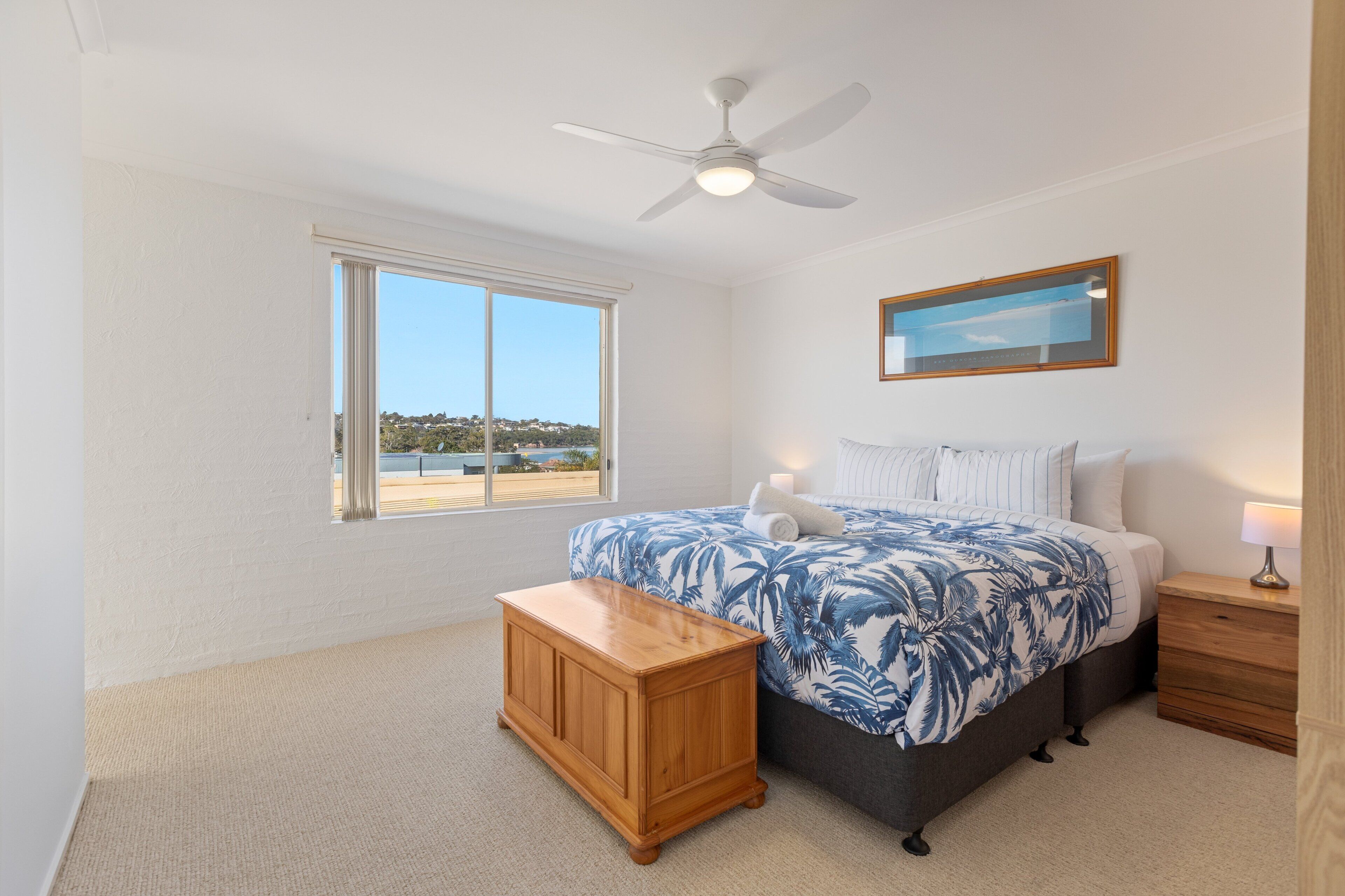A bedroom with a bed, a nightstand, a wooden bench, a ceiling fan, a window with a view, and a framed picture on the wall.