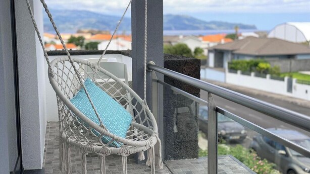 Een balkon met een macramé schommelstoel, een glazen balustrade en uitzicht op gebouwen en bergen.