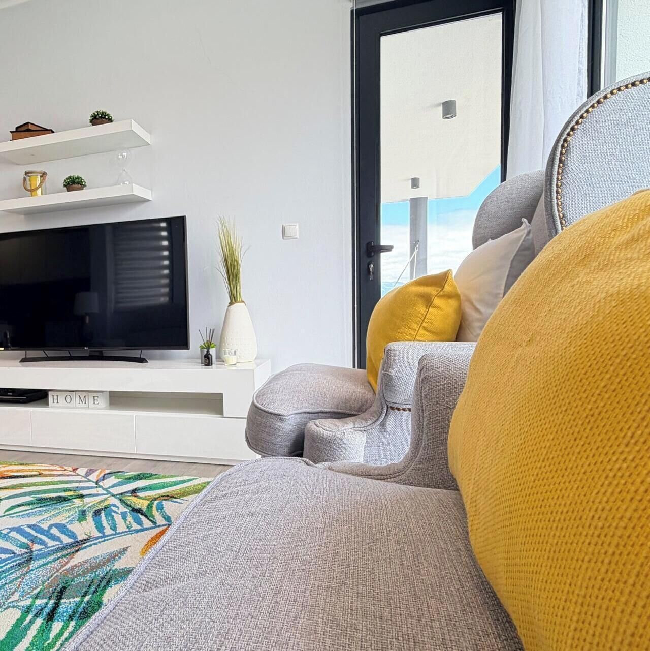 A modern living room with a flat-screen TV, a sofa with a mustard-colored cushion, and a decorative rug.