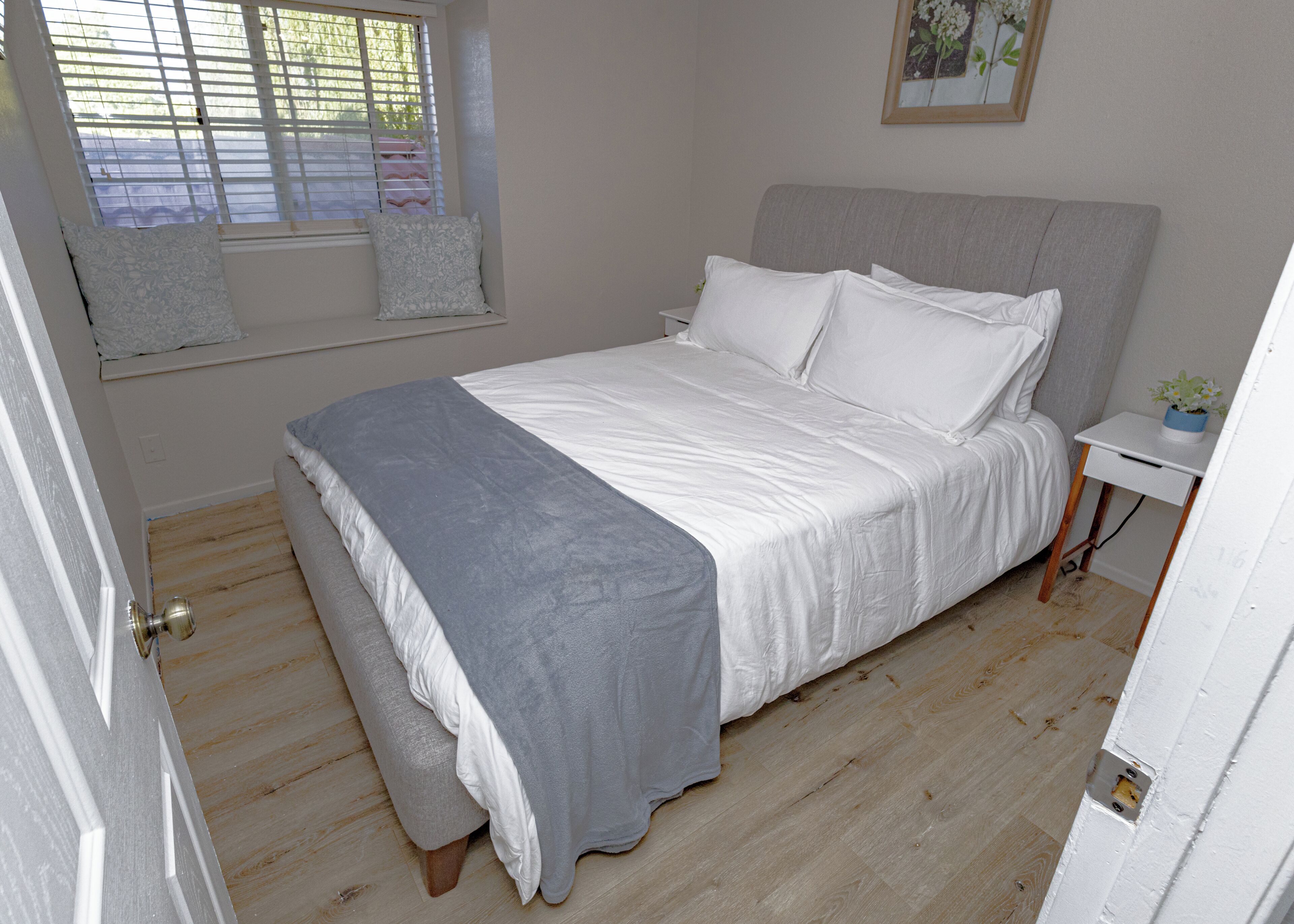 Ein Schlafzimmer mit einem Bett, einem Nachttisch mit Pflanze, einem Fenster mit Jalousien und einer Tür.